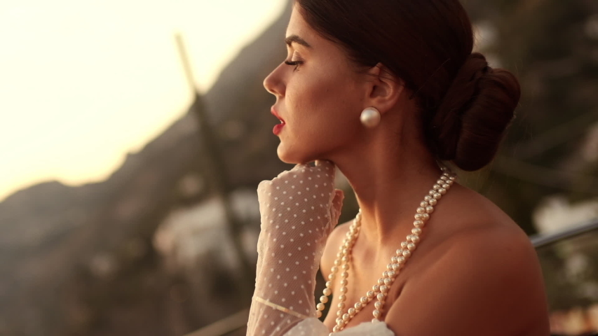 Portrait Of Pretty Brunette In Elegant White Dress. Girl In Jewelry With Pearls And White Gloves Posing On The Street In Positano. Italy.