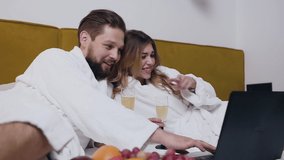 Lovely joyful amorous 30s couple dressed in white bathrobes resting on comfortable hotel bed and holding in their hands glasses with champagne enjoying revision of amusing videos on laptop