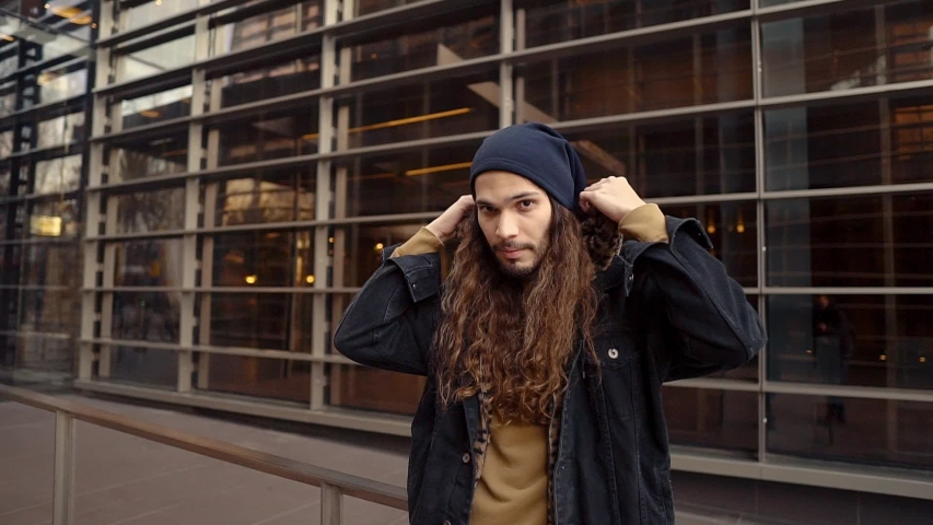 Portrait of long haired hipster walking on street in modern city in slow motion. He wears black jacket with leopard hoodie. Stylish man near business buildings
