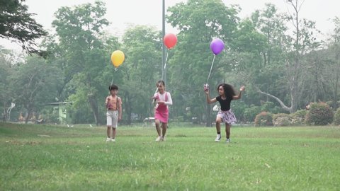 Slow motion cheerful group of little cute kids running on the green grass competitive together with multicolored balloon in the public park. Friendship togetherness concept.