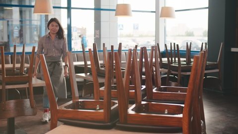 Portrait of young asian waitress or owner in uniform putting chairs on floor from tables preparing cafe before open it. Conception of small business, startup and food service วิดีโอสต็อก