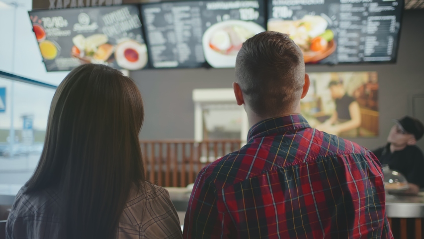 Back view of young customer guy and girl look at menu on wall in coffee shop choose drink, man and woman pointing fingers at menu. couple cafe chooses dish and drinks to order. Royalty-Free Stock Footage #1045390888
