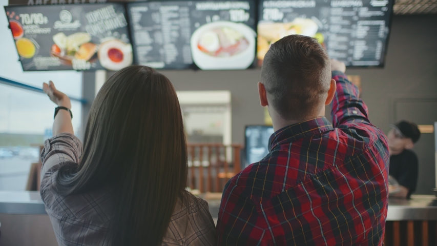 Back view of young customer guy and girl look at menu on wall in coffee shop choose drink, man and woman pointing fingers at menu. couple cafe chooses dish and drinks to order. Royalty-Free Stock Footage #1045390888