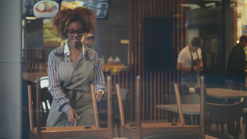 Portrait of beautiful afro-american waitress in apron putting chairs on table preparing cafe for opening. Waiting staff cleaning restaurant. Small business and food service concept. Royalty-Free Stock Footage #1045390894