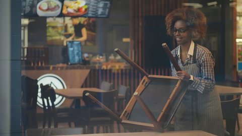 Portrait of beautiful afro-american waitress in apron putting chairs on table preparing cafe for opening. Waiting staff cleaning restaurant. Small business and food service concept.: film stockowy