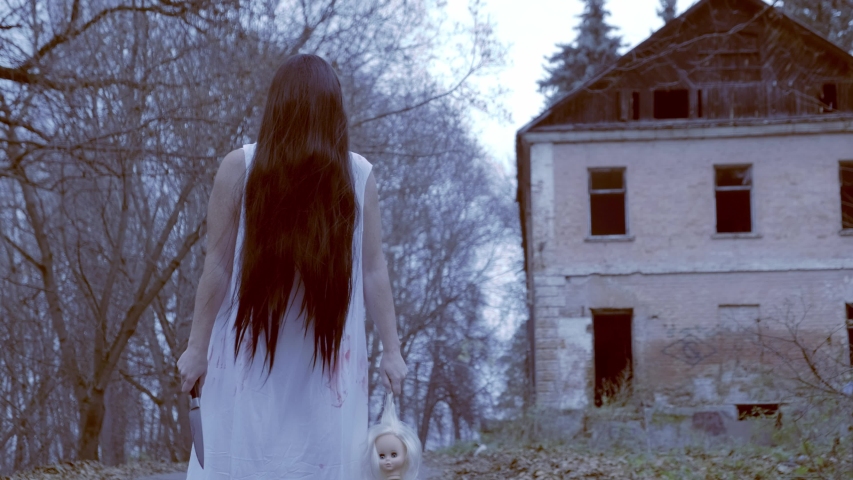 Mystical girl with black long hair on background of ominous house. ghost of dead girl holds doll’s head and knife in her hands.