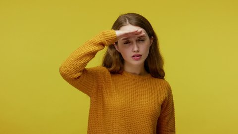 Curious girl in casual clothes holding hand over eyes and looking far away with attentive view, ambitious student making plans for future, watching perspectives. indoor studio shot yellow background