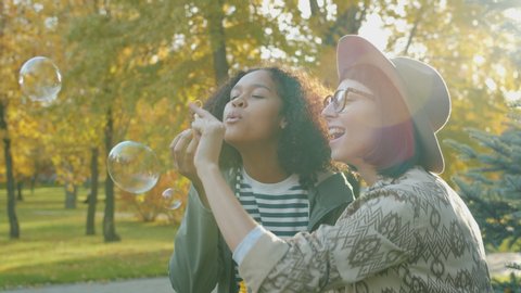 Portrait of happy girls friends blowing soap bubbles in park in autumn laughing having fun enjoying funny activity. Happiness and friendship concept.