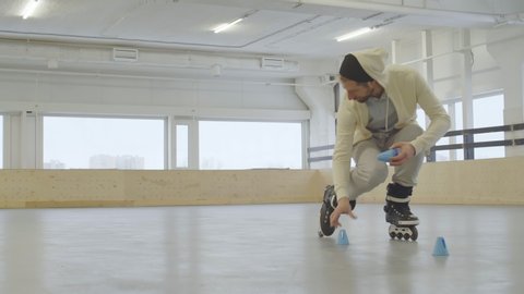 Wide shot of Caucasian male inline skater wearing professional roller-skates skating backwards and putting blue cones on floor