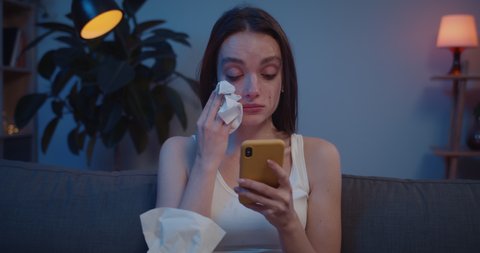 Close up of young woman wiping tears from her face with paper tissue while looking at phone screen. Upset girl cant believe bad news reading on her smartphone and crying.