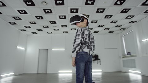 Tracking arc 360 degree shot of a little boy is looking around in virtual reality wearing headset in a big empty white room – Stockvideo