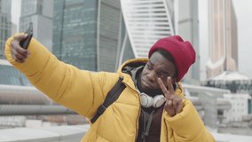 Medium shot of young African man wearing yellow jacket, red hat and white headphones standing in city center and making selfie on telephone