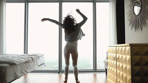 Back rear view attractive young woman dancing in front of huge window, listening to favorite music at home. Smiling lady having fun in bedroom, jumping twisting, feeling joyful alone, slow motion.