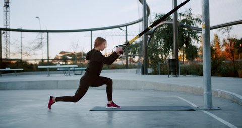 Side shot of strong Caucasian female doing leg swing using TRX suspension straps outdoor, diligent fitness workout of attractive girl in black leggings and hoodie, sportive lifestyle urban landscape Stockvideo