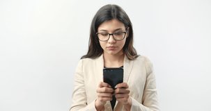 Attractive dark haired woman in business outfit and protective eyeglasses texting message on black smartphone while standing over white studio background. Young lady using modern technology at work.