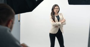 Attractive brunette woman in formal clothing and eyeglasses standing with crossed hands over white background while specialist taking her pictures at modern studio. Concept of shooting process.