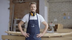Portrait video of confident middle aged carpenter in apron looking at camera posing with his hands on hips in woodworking shop, unfinished wooden boat model placed on workbench behind him