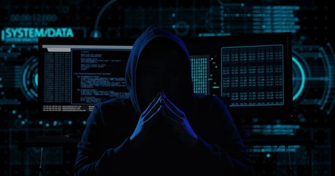 Portrait of mysterious hooded hacker sitting at desk and looking at the camera after breaks into government data servers in dark atmosphere with system codes animation in background.