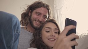 Close up view of joyful lovely couple using smartphone while sitting together near the bed at home