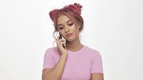 A displeased young woman with the color pink hairstyle is hiding her discontent while talking on the phone over white background