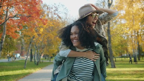 Slow motion of happy girl giving piggyback ride to joyful lady friend in park on sunny autumn day. Happiness, friendship and modern lifestyle concept.