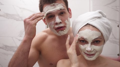 Happy positive young couple man and woman having fun while standing in a bathroom after shower. Both playfully applying white mask on face. Having fun. Portrait couple together applying face mask