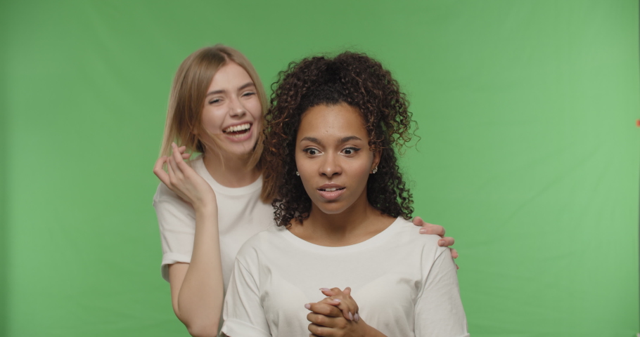Two women gossiping, teenage girl sharing secret information with shocked surprised friend, whispering to ear, Female having fun on a Green Screen croma key, 4k raw video footage slow motion 60 fps