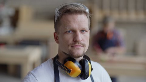 Head and shoulders video portrait of serious middle aged carpenter looking at camera in carpentry shop with protective earmuffs around his neck while smb woodworking in background, copy space to right