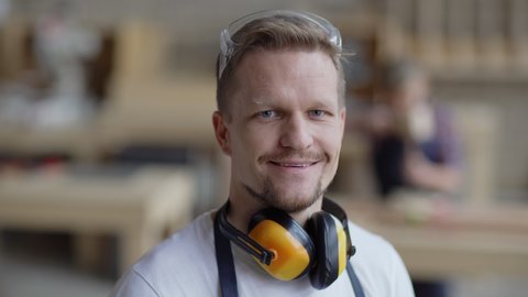 Head and shoulders portrait of middle aged carpenter looking at camera and smiling in carpentry shop with protective earmuffs around his neck while smb working in background, copy space to right