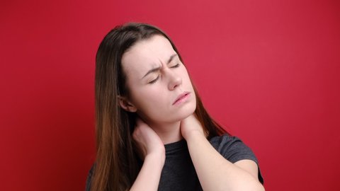 Tired upset young woman feeling stiff sore neck pain concept rubbing massaging tensed muscles suffer from fibromyalgia, has gloomy expression, isolated over red background. Fibromyalgia concept