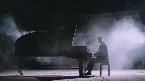 Professional pianist in dark suit playing on a grand piano on big stage in concert hall with dimmed light in the smoke