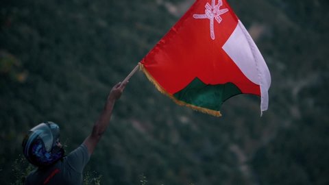  Shaat, Dhofar/Oman - November  17, 2019 :
shot of a young Omani man holds the flag of the Sultanate of Oman over the top of the mountain during sunset in the south of Sultanate of Oman .