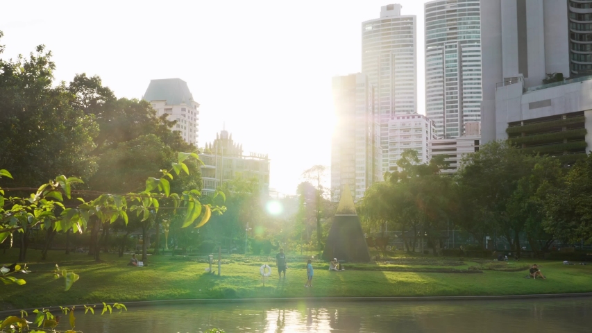 People Relax in Public Park with Lake and Green Trees with Skyscrapers Buildings in the City on Background and Bright Sunshine of Sunset. Park for Rest, Leisure and Fun in Center of Modern Metropolis | Shutterstock HD Video #1045525930