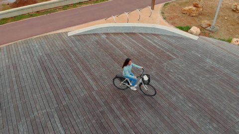 In the background, a girl rides a Bicycle on a round stadium, top view. She's wearing a blue jacket and jeans. It is located near the sea.