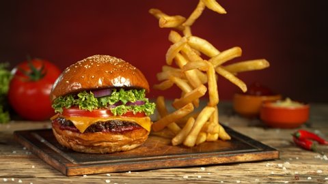 Super Slow Motion Shot of Fast Food Concept. Falling French Fries on Wooder Cutting Board next to the Fresh Hamburger at 1000fps.