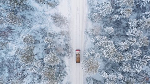 Drone view of red truck with cargo on snow covered empty road among winter snowy forest