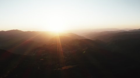 Lens flare in mountains at orange sunset in summer evening aerial view drone. Sun rays. Green pine forest slopes of mountain range. Bright disk of sun in blue sky slowly sets tops of mountains. Nature