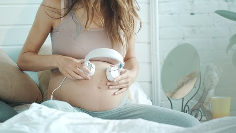 Macro midsection pregnant woman applying headphones on her belly in bed. Expectant mother listening music in light bedroom. Pregnant female giving unborn listening music at home.