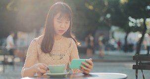 Smiling asian girl watching videos on smartphone and drinking coffee outdoors. Young student girl having coffee breack and using her smartphone. City background.Concept of happy lifestyle
