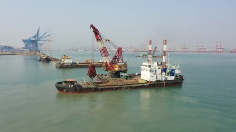 QINGDAO, CHINA – SEPTEMBER 2019: Rotating aerial view of excavating machinery dredging sand and other seabed material, scooping it onto industrial boat near construction site in Port of Qingdao, China