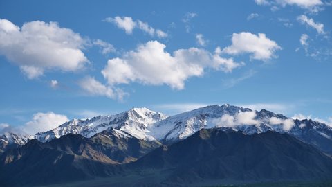 Time lapse of clouds and snow clad mountains while blue sky in the background. Shadow of clouds creating patterns on the mighty barren mountains