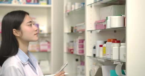 Young asian pharmacist woman holding computer tablet using for checking medication details on a box in pharmacy drugstore.