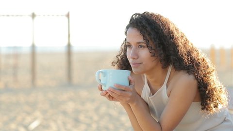 Happy latin woman drinking coffee on the beach at sunset enjoying flavor in slow motion