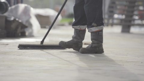 Construction worker cleans a building site with a industrial vacuum cleaner. Man uses broom in angled sunlight to sweep towards ground based camera and then past.
