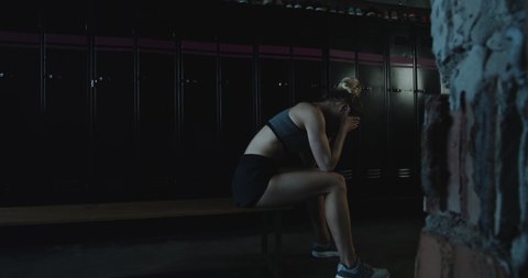 Upset athletic blonde woman sitting alone, lying down on bench in dark gym locker room tired after bad day slow motion.