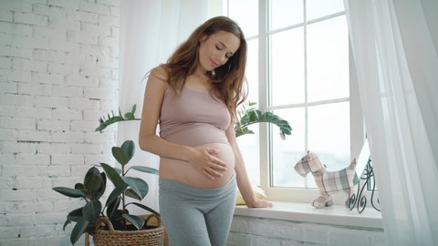 Happy pregnant woman stroking belly near window indoors. Pretty expectant mother standing in light room. Future mom looking on window in slow motion. Pregnant woman smiling to unborn baby