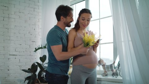 Husband giving yellow flowers to pregnant wife indoors. Pregnant couple hugging near window at home. Expectant mother chatting mobile phone in slow motion. Surprised pregnant woman