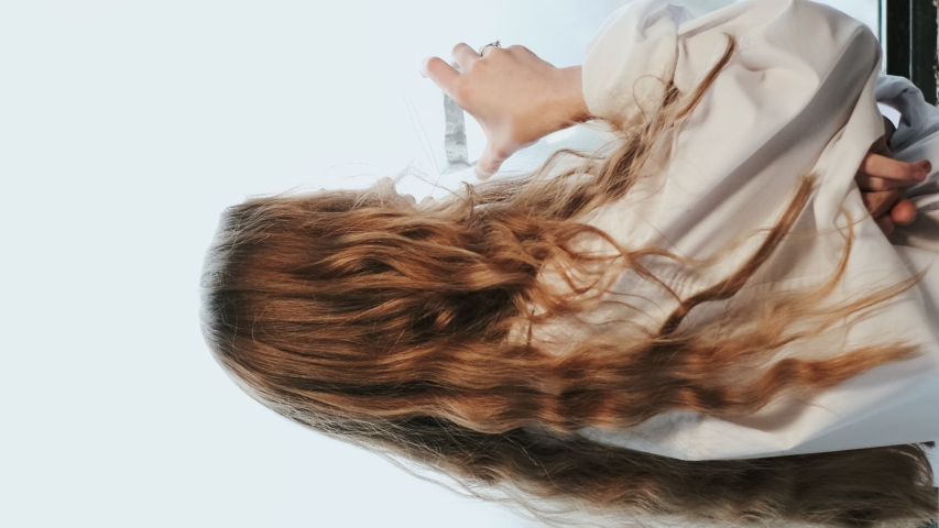 Young Girl With Long Hair Stands By The Window With Glass Of Water. Seductive Redheaded Girl. Girl In White Shirt On Her Shoulders Falling Curls.