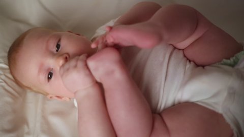 The child is trying to eat his legs. Little cute girl lies on her back in a white bodysuit and puts her feet in her mouth.