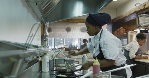 Side view of an African American female cook working in a busy restaurant kitchen, cooking in a frying pan, her colleagues working in the background